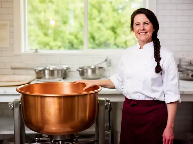 Smiling woman with a dark braid in a white chef jacket resting her hand on a large copper kettle